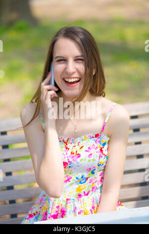 Portrait of young beautiful woman with long hair in summer park, girl wearing flower short dress is using a smartphone while sitting on a bench Stock Photo