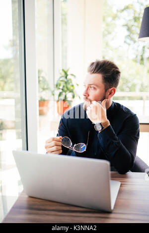 bearded businessman glasses in hand and sitting at his laptop and look in the window. Stock Photo