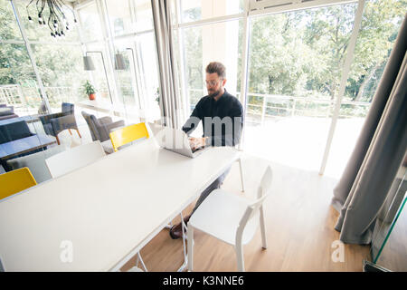 Bearded businessman sitting at the big conference romm white table and working at laptop before meeting Stock Photo