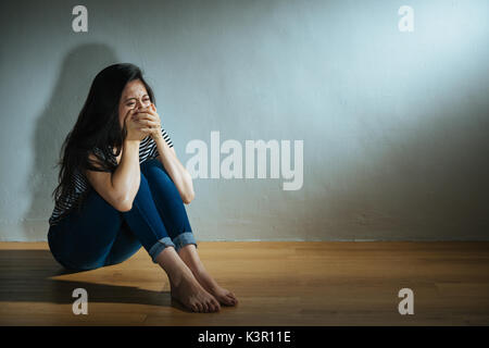 battered abused women concept of depressed woman sitting on wood floor in dark white wall room crying and hand cover mouth sadness feeling afraid. Stock Photo