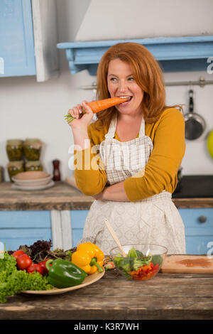 Portrait of housewife standing in the kitchen, biting carrot. Stock Photo