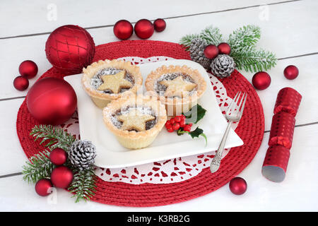 Christmas mince pies with holly on a porcelain plate with holly, silver fork, fir, cracker and red bauble decorations on distressed white wood backgro Stock Photo