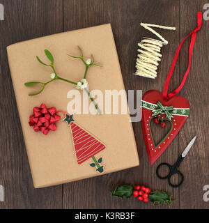 Christmas gift wrapping background with parcel, tag, ribbon, scissors, heart shaped bauble, holly, mistletoe and string on oak wood. Top view of xmas  Stock Photo