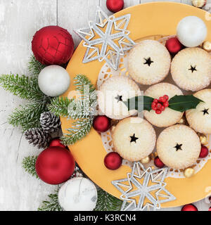 Christmas mince pies on a gold plate with holly, fir, bauble decorations on distressed wood background Stock Photo