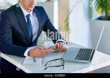 Portrait of successful young businessman using smartphone while working at small table in cafe during coffee break Stock Photo