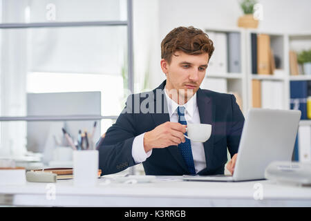 Portrait of modern businessman working in office drinking coffee at desk and using laptop Stock Photo