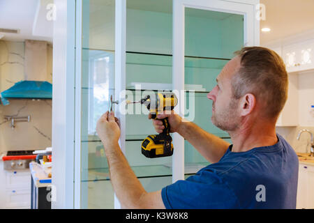 The worker sets a new handle on the white cabinet with a screwdriver installing kitchen cabinets Stock Photo