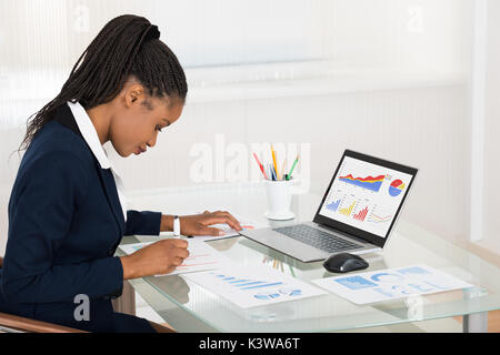 Young African Businesswoman Analyzing Graph While Laptop On Office Desk Stock Photo