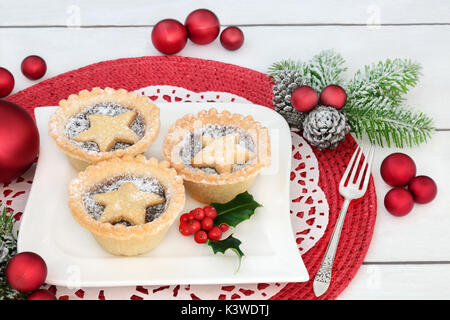 Mince pies with holly on a porcelain plate with old silver fork, fir and bauble decorations on red place mat on distressed white wood background. Stock Photo