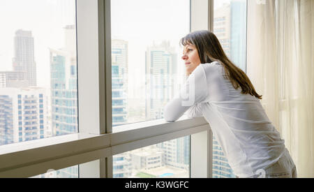 Mature woman is looking out of the window in her hotel/apartment Stock Photo