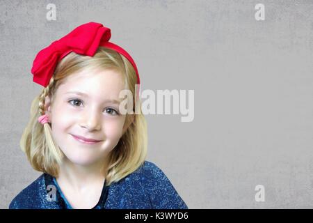 Portrait of a little six years old smiling cute blond girl with a red bow on his head on gray grunge background. Stock Photo