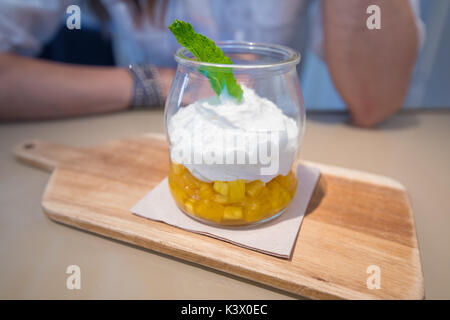 white yogurt with pineapple chunks and mint leaf in glass jar on wooden plank, next to woman ready to eat Stock Photo
