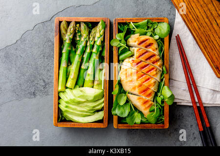 Healthy lunch in wooden japanese bento box. Balanced healthy food grilled chucken and avocado with asparagus and green salad. Top view, slate gray bac Stock Photo