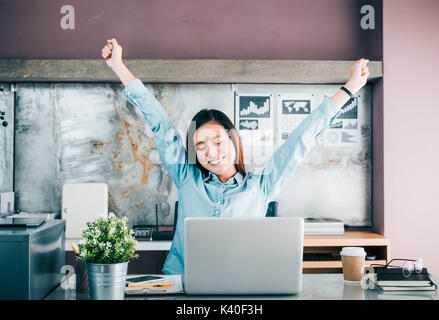 Asian creative designer raise arm up and close eye in front of laptop computer on desk,relax from hard work in office. Stock Photo
