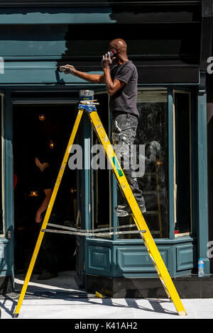 Painter and Decorator Using Mobile Phone (Cell Phone) Whilst Painting A Shop Front, London, UK Stock Photo