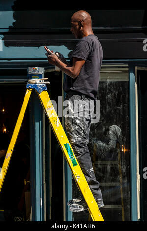 A Painter and Decorator Takes A Break From Painting A Shop Front To Answer A Telephone Call, London, UK Stock Photo