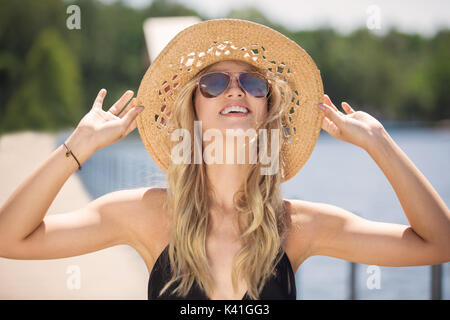 A photo of beautiful woman in bikini and panama hat standing on the pier and smiling happily. Stock Photo