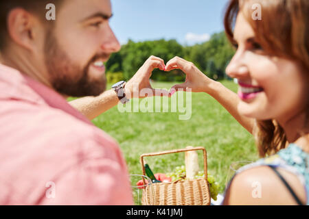 A photo of young, happy couple celebrating their anniversary on the picnic at the park. They're putting their hands together in the heart shape. Stock Photo