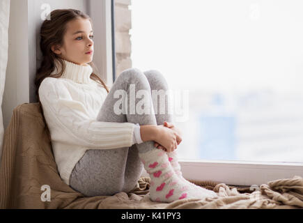 sad girl sitting on sill at home window in winter Stock Photo