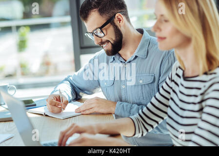 Smiling man writing in notebook while colleague working on laptop Stock Photo