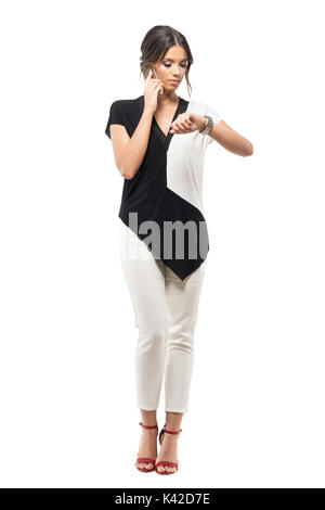 Busy business woman checking time on wrist watch talking on the mobile phone. Full body length portrait isolated on white studio background Stock Photo