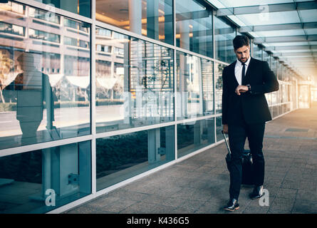 Businessman carrying a suitcase and looking at his watch while walking in airport. Business traveler checking time on his wristwatch. Stock Photo
