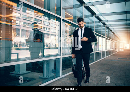 Businessman walking with luggage and using mobile phone at airport. Young man on business trip text messaging from his cell phone. Stock Photo