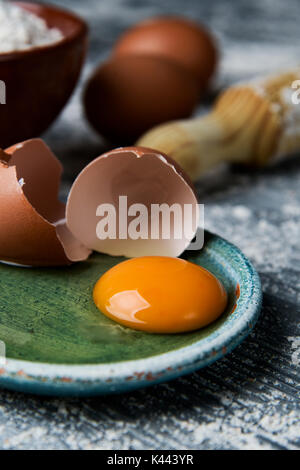 closeup of a cracked egg in a green plate, an earthenware bowl with flour, some whole eggs and a wooden rolling pin on a rustic table sprinkled with f Stock Photo