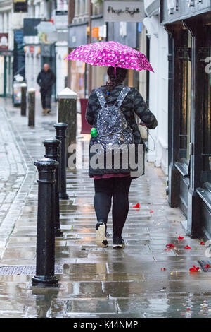 Woman walking holding a pink umbrella above her head to prevent rain falling on her along pavement in Chester City, Cheshire in pouring rain Stock Photo