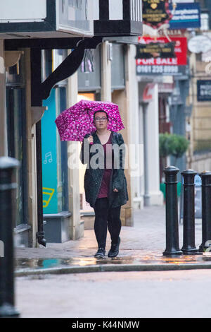 Woman walking holding a pink umbrella above her head to prevent rain falling on her along pavement in Chester City, Cheshire in pouring rain Stock Photo