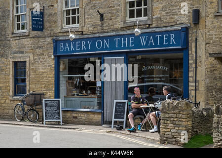 Cycling tourists enjoying the Summer Sun, a coffee & a snack outside an artisan bakery in the historic Cotswold village of Bourton on the Water Stock Photo