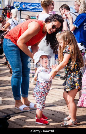 Little girl, 4-5 years, helping her baby sister to stand outside in sunshine on wooden deck, Mother bends to watch, smiling. Other people around. Stock Photo