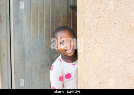 Young Ugandan girl dressed in bed clothing leans around the door and smiles at the camera in the rural countryside in Uganda. Stock Photo