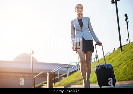 Bottom view on mature glad businesswoman walking and pulling suitase. Stock Photo
