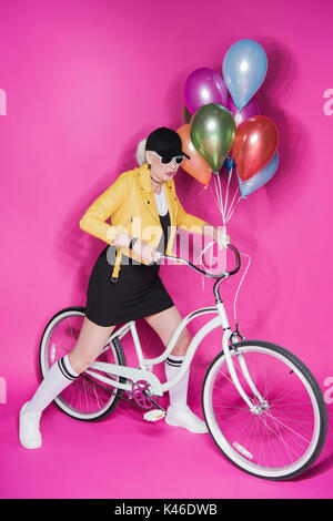 fashionable senior woman wearing yellow leather jacket holding colorful balloons and riding bicycle isolated on pink Stock Photo