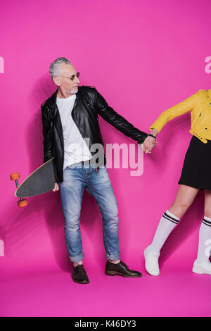 Stylish senior couple in leather jackets posing with skateboard and holding hands isolated on pink Stock Photo