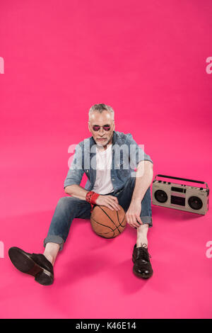 senior man sitting on floor with tape recorder and basketball ball isolated on pink Stock Photo