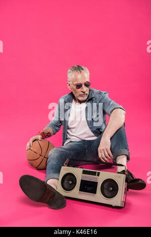 senior man sitting on floor with tape recorder and basketball ball isolated on pink Stock Photo