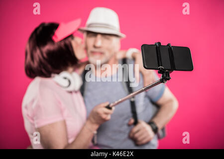selective focus of senior woman kissing husband while taking selfie isolated on pink Stock Photo