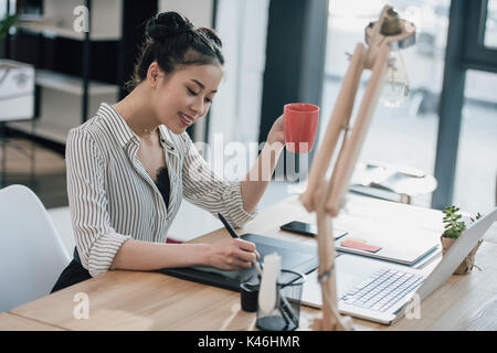 Smiling young asian businesswoman using graphic tablet and drinking coffee in office Stock Photo