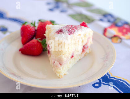 Strawberry cake and three strawberries with white cream on top, soft focus, closeup, on white porcelain plate, on top of a blue cloth on kitchen table Stock Photo