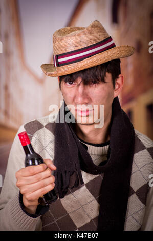 Close up of a sad homeless young man in the streets, wearing a hat and a scarf, and holding a bottle of wine in his hand, in a blurred background Stock Photo