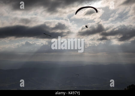 Some para-gliders and hang-gliders flying over a valley, with distant mountains and hills and sun rays coming out through the clouds Stock Photo