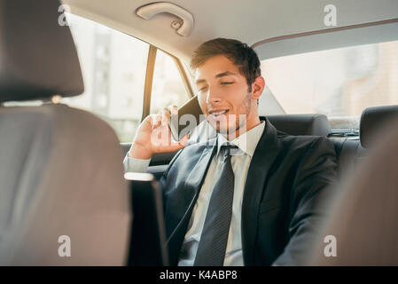 Businessman working while sitting in a car. Man beeing driven to work in his limo. Suit and tie businessman in the back seat making a call while the limo driver is driving. Stock Photo