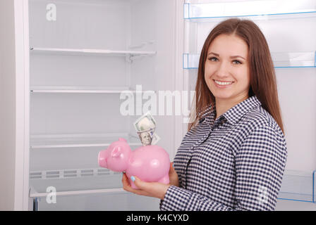 Young beautiful woman standing with piggy bank (money box) on the refrigerator background Stock Photo