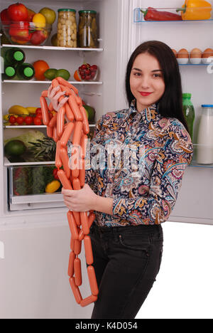 Young girl holds sausages on the refrigerator background. Beautiful young girl near the fridge. Stock Photo