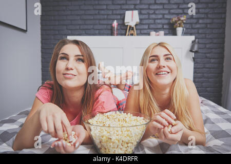 Portrait of two happy women watching movie on bed eating popcorn. Stock Photo