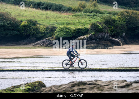 Gannel Estuary - A man riding a mountain bike crossing over the Gannel River via a footbridge at low tide. Stock Photo