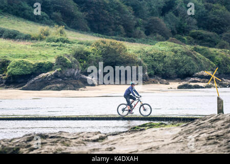 Gannel Estuary - A man riding a mountain bike crossing over the Gannel River via a footbridge at low tide. Stock Photo