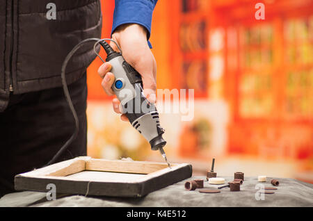 Closeup of a hardworker man drilling a wooden frame with his drill, with drilling accessories over a gray table in a blurred background Stock Photo